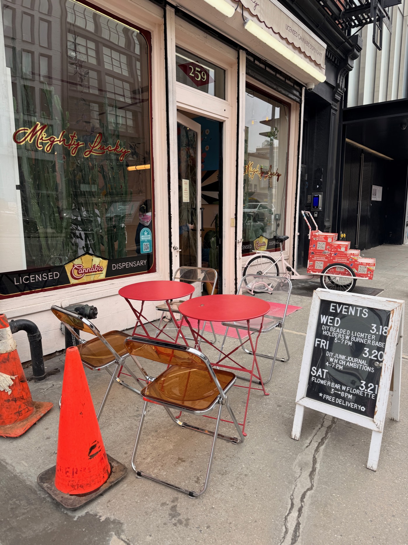 Mighty Lucky storefront at 259 Bowery — gold script neon in the window, Licensed Cannabis Dispensary awning, red cafe tables and a pink cruiser bike on the sidewalk